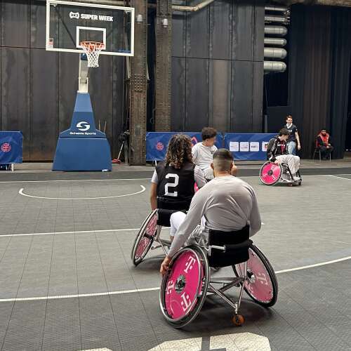 Mehrere Personen sitzen in Rollstühlen. Im Hintergrund steht ein Basketballkorb.