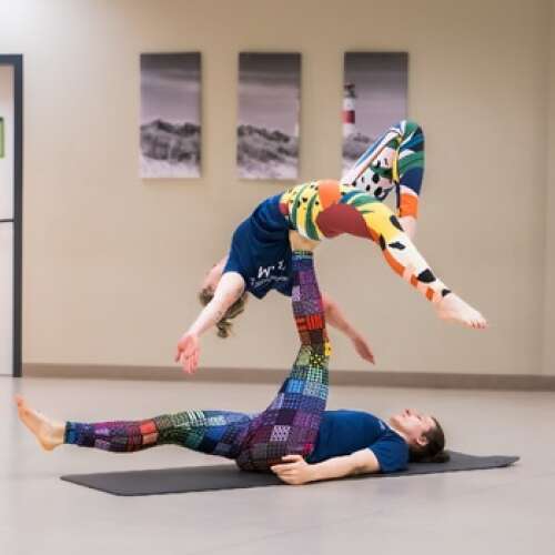 Eine Person liegt in Rückenlage und trägt auf einem nach oben ausgestrecktem Bein eine andere Person. Diese liegt mit ihrem Rücken auf dem Fuss der unteren Person. Sie streckt ihre Arme zur Seite; ein Bein ist angewinckelt und das andere Bein ist gestreckt. 