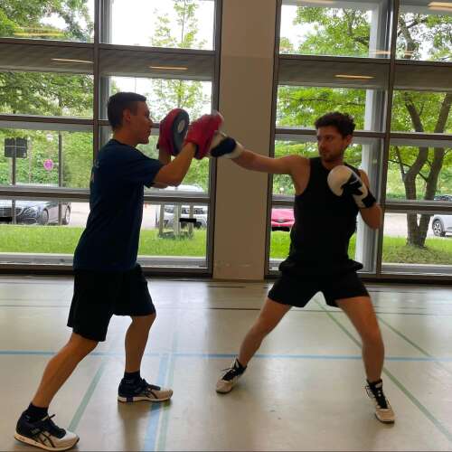Auf dem Bild stehen sich zwei Personen gegenüber, die eine Übung mit Boxhandschuhen und Handpratzen durchführen. Eine Person boxt mit der hinteren linken hand in die Pratze seines Gegenübers.