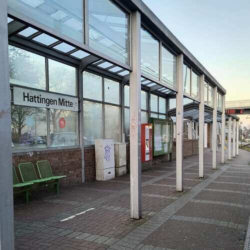 Das Bild zeigt die Haltestelle Hattingen (Ruhr) Mitte. Zu erkennen ist die überdachte Haltestelle mit einer grünen Bank, dem roten Ticketautomaten und dem Haltestellenschild.