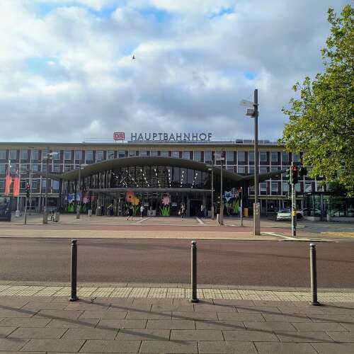 Das Bild zeigt den Eingangsbereich eines modernen Hauptbahnhofs mit dem gut sichtbaren „DB Hauptbahnhof“-Schriftzug. Vor dem Gebäude verlaufen eine breite Straße und ein Platz, während der Himmel von großen Wolken geprägt ist und die Szene freundlich beleuchtet.