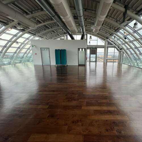 Das Bild zeigt einen großzügigen, lichtdurchfluteten Raum mit geschwungener Glasdecke, sichtbaren Lüftungsrohren und dunklem Holzboden. Er wirkt modern und offen, während in der Mitte ein weißer Kubus mit Türen und zwei türkisfarbenen Matten steht.