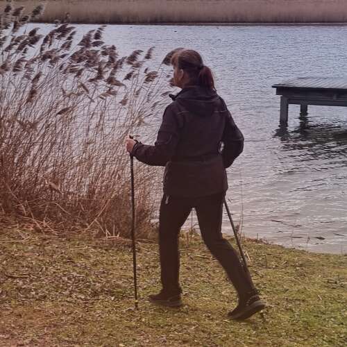 Das Bild zeigt eine Person, die zwei Nordic Walking Stöcke in der Hand hat und über eine Wiese läuft. Im Hintergrund sieht man einen See und das Ende eines Steges.