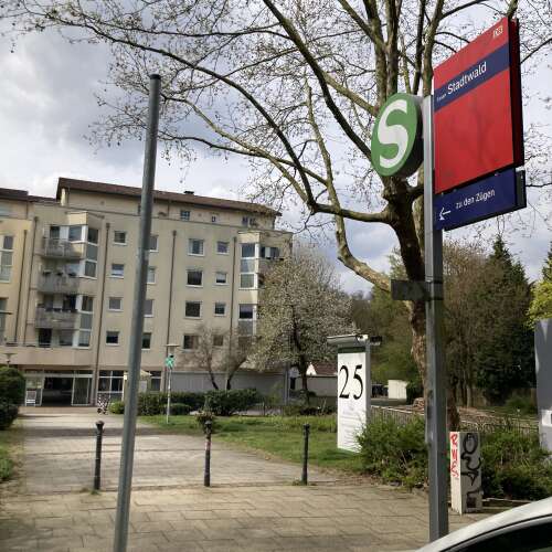 Ein S-Bahn-Schild mit der Aufschrift „Essen Stadtwald“ steht neben einem Baum an einem ruhigen Weg. Im Hintergrund sind ein Wohngebäude, blühende Bäume und Grünflächen zu sehen.