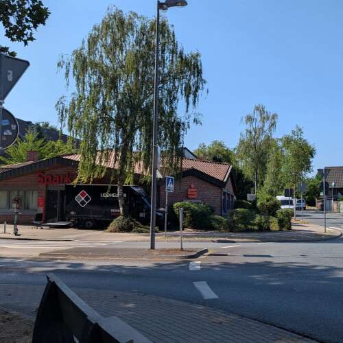 Das Bild zeigt den Haupteingang der Sparkasse Bochum, Geschäftsstelle Grumme, an einer Straßenecke mit Zebrastreifen und Kreisverkehr. Der Eingang ist durch das rote Sparkassen-Logo und den Schriftzug gut erkennbar.