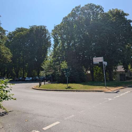 Das Bild zeigt einen Kreisverkehr mit einer begrünten Mittelinsel, Wegweisern und großen Bäumen. Der Treffpunkt befindet sich an der Straßenecke in unmittelbarer Nähe der Beschilderung.