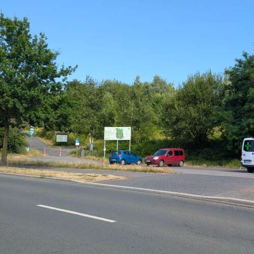 Ein Weg führt von einer Straße in ein bewaldetes Naherholungsgebiet, daneben befinden sich Informationstafeln und einige parkende Autos.