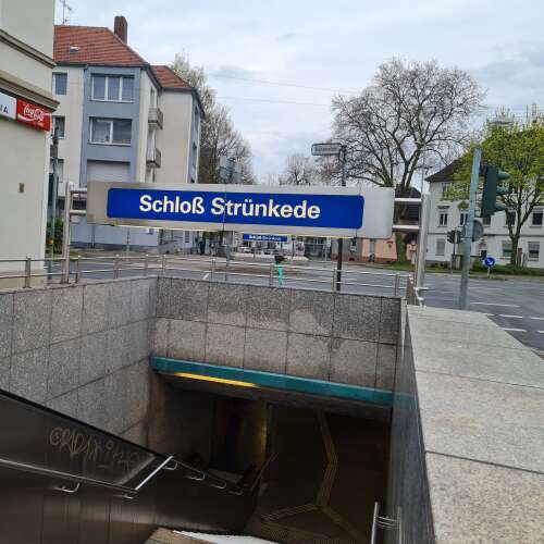 Ein U-Bahn- bzw. Unterführungseingang mit dem Schild „Schloß Strünkede“ befindet sich an einer ruhigen Straßenkreuzung. Im Hintergrund stehen Wohnhäuser, Bäume ohne Blätter und eine Verkehrsampel unter einem grauen Himmel.